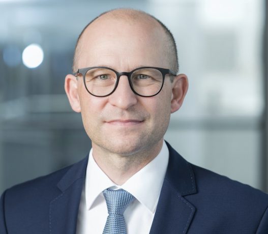 Ein Mann in einem marineblauen Anzug, einer blau gemusterten Krawatte und einer Brille lächelt leicht. Er hat kurzes, helles Haar und steht vor einem verschwommenen Bürohintergrund, der mit kühlen Stahltönen akzentuiert ist.